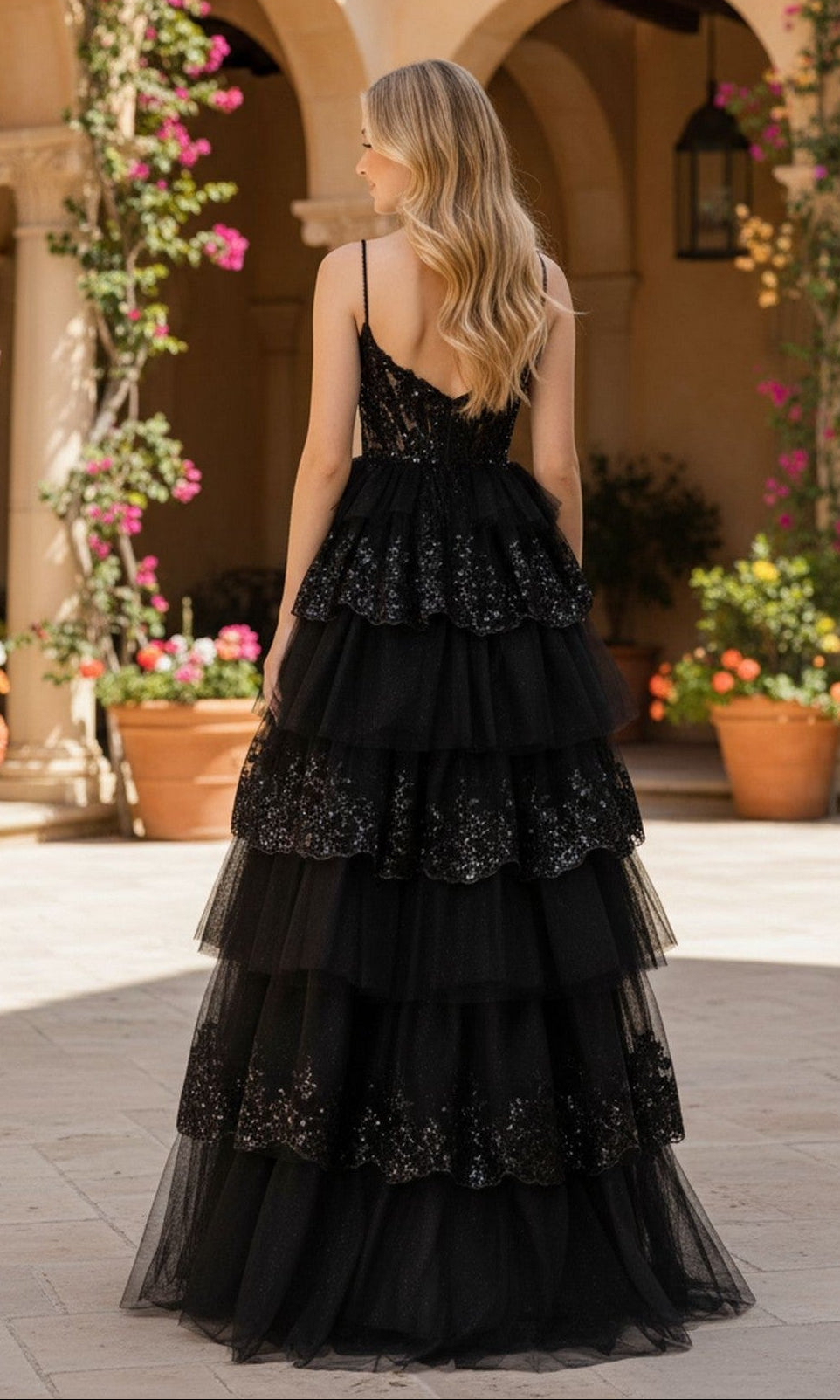 A woman with long wavy blonde hair wears the Sparkly Prom Ball Gown: PromGirl Exclusive 250074, posing outdoors in a courtyard with potted plants and arched architecture.